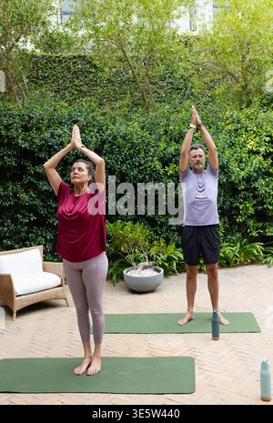 Coppie anziane diverse che fanno yoga in piedi su tappeti verdi sul patio indossando un top Bordeaux con bottiglie Foto Stock
