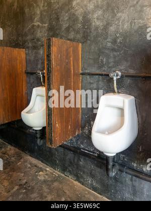 Elegante bagno maschile caratterizzato da orinatoi bianchi contro una parete di cemento con loft scuro e divisori in legno rustico in un ristorante o un bar interno moderno. Foto Stock