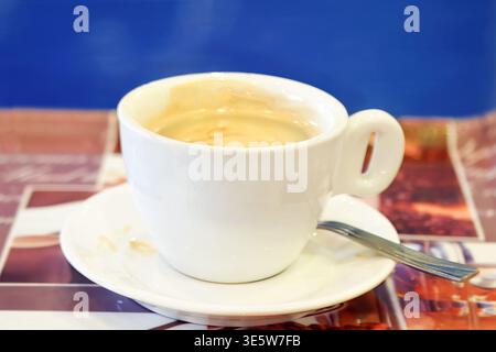 Una tazza bianca riempita di espresso cremoso si trova su un piattino, con un cucchiaio accanto. Lo sfondo blu intenso contrasta le tonalità calde del caffè. Foto Stock