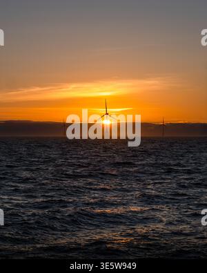 Una fila di turbine eoliche offshore si staglia contro un vivace tramonto arancione e dorato sul Mare del Nord, con colline costiere visibili oltre. Foto Stock