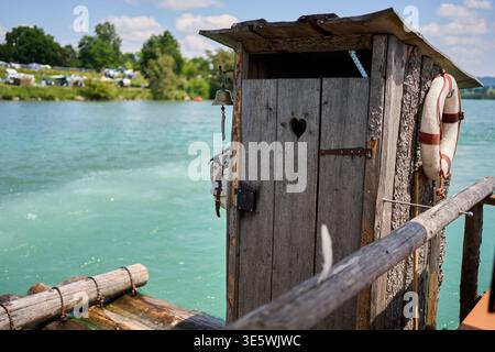 Toilette rustica in legno dell'attico su una zattera che galleggia su un fiume. Presenta un ritaglio a forma di cuore, un salvagente e una campana, con un paesaggio estivo alle spalle. Foto Stock