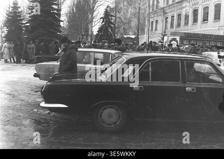 Sloviansk, fine anni '1980, fabbrica AIZ. Un'impressionante fotografia d'archivio in bianco e nero che cattura il corteo ufficiale di una delegazione di alto livello. Diverse classiche auto executive sovietiche, tra cui la Volgas nera, sono parcheggiate sulla strada innevata. Sullo sfondo, una grande folla di lavoratori locali e cittadini in abbigliamento invernale si è riunita per assistere alla visita storica. Questa affascinante scena documentaria riflette perfettamente gli eventi pubblici, la vita urbana quotidiana prima della guerra e un Donba tranquillo, unito e pacifico. Foto Stock