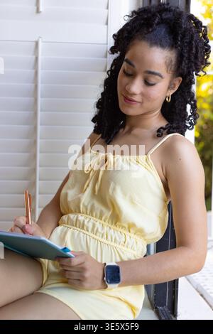 Donna in romper giallo pallido seduta sul bordo di persiane bianche che scrivono su un notebook con penna Foto Stock