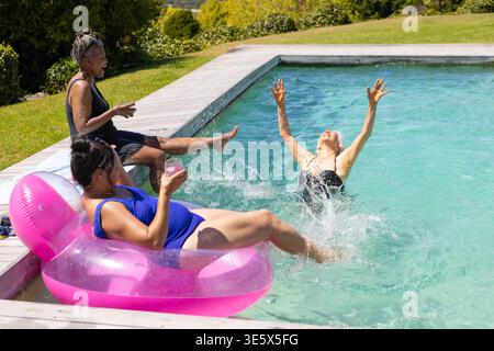 Diverse donne che si rilassano in costume da bagno su un galleggiante rosa nella piscina sul cortile con vetro trasparente e spruzzi Foto Stock