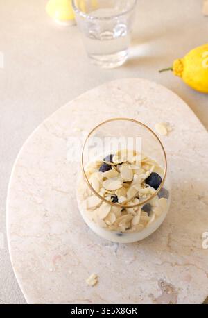 Parfait di yogurt sano con fiocchi di mandorle e mirtilli in vetro su vassoio di marmo Il primo piano include limone fresco e acqua sullo sfondo Foto Stock