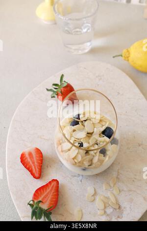Parfait di yogurt sano con fiocchi di mandorle mirtilli e fragole su vassoio di marmo La foto ravvicinata è dotata di recipiente in vetro con limone fresco e acqua Foto Stock