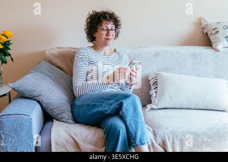 Donna ricciosa a 40 anni che usa lo smartphone seduto sul divano di casa Foto Stock