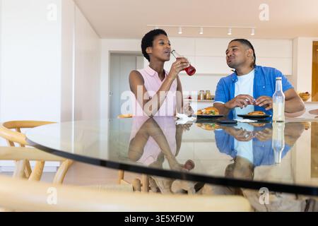 Parlare e sorseggiare diversi amici in cucina al tavolo di vetro, con panini e bottiglia di vetro Foto Stock