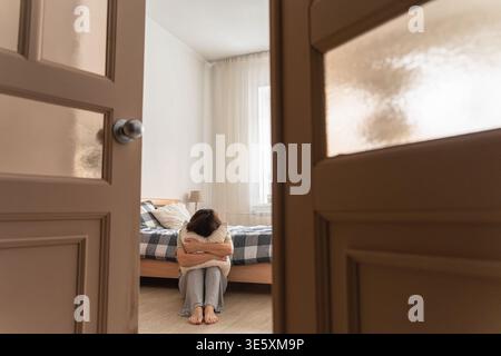 Donna che abbraccia il cuscino mentre siede sul pavimento della camera da letto, si sente sola, triste e soffre di problemi di salute mentale Foto Stock