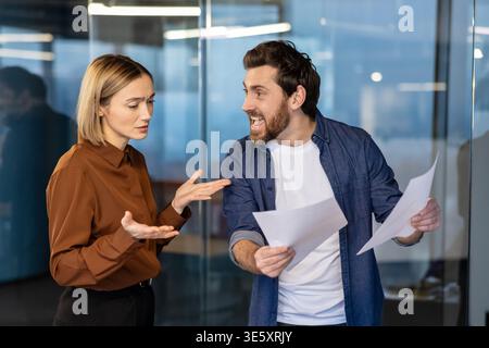 Uomo con documenti in mano e urla contro una donna con espressioni frustrate e mani aperte, che raffigura un acceso litigio e un conflitto di lavoro in un ambiente di ufficio moderno Foto Stock