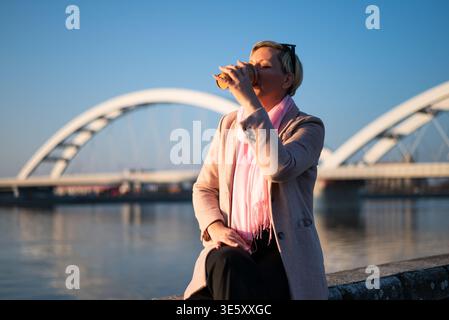 Donna urbana che si rilassa all'aperto vicino al fiume, bevendo caffè caldo. Giorno di sole, ponte cittadino sullo sfondo, momento di vita tranquillo. Foto Stock