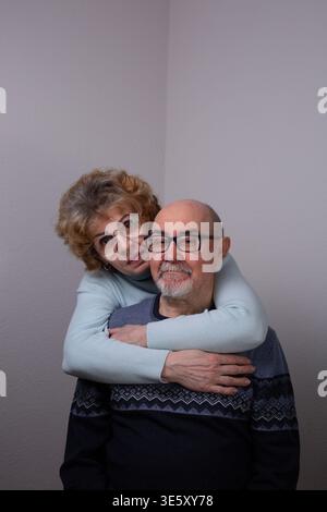 Una donna anziana felice che abbraccia il marito da dietro. Uomo e donna maturi che posano insieme in casa, esprimendo amore, sostegno e relazioni a lungo termine Foto Stock
