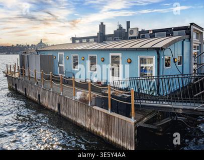 Un moderno edificio galleggiante blu con un ponte in legno e luci in stile oblò si trova sul tranquillo lungomare di Stoccolma. Foto Stock