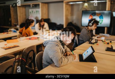Giovane studente che studia con un tablet e prende appunti in una classe inclusiva con colleghi Foto Stock