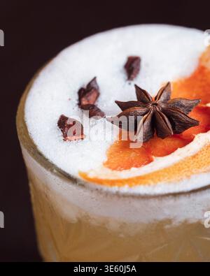 Bevanda schiumosa con anice stellato e fetta di arancia. Il ricco liquido ambrato suggerisce un cocktail caldo e aromatico. Lo sfondo scuro migliora il vivace colon della bevanda Foto Stock