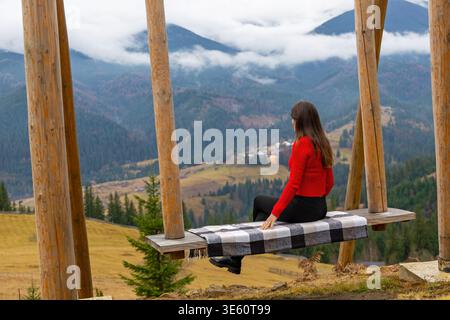 Donna sull'altalena che si affaccia sulla Cloudy Mountain Valley. Una donna con maglione rosso siede su un'altalena di legno, ammirando una valle nebbiosa di montagna e boscosa Foto Stock