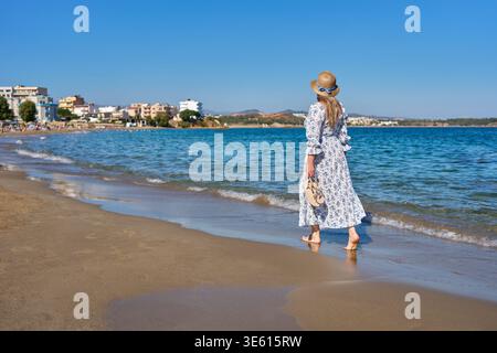 Donna con maxi abito floreale e cappello di paglia che cammina a piedi nudi lungo una spiaggia sabbiosa a Chania, Creta. Splendido mare blu e sfondo della costa della città. Foto Stock