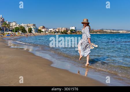 Donna felice in un maxi abito floreale e cappello di paglia che cammina a piedi nudi lungo la spiaggia di Nea Chora a Chania, Creta. Soleggiata giornata estiva sul Mar Mediterraneo. Foto Stock