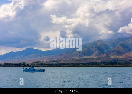 La barca passeggeri blu trasporta diversi gruppi di persone attraverso il tranquillo lago Issyk-Kul in Kirghizistan. Montagne maestose e piccola città creano un'atmosfera suggestiva Foto Stock