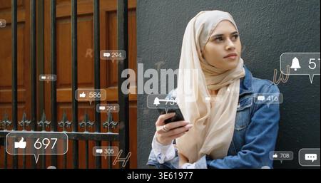 Donna pendente che indossa hijab beige e giacca in denim che tiene il telefono al cancello con avvisi e spazio per le copie Foto Stock