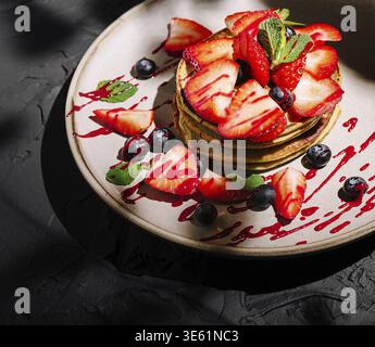 Il cibo, i pancake alti sono impilati artisticamente su un piatto, adornati con fragole a fette, mirtilli e un filo di salsa di bacche, adornati con un da Foto Stock