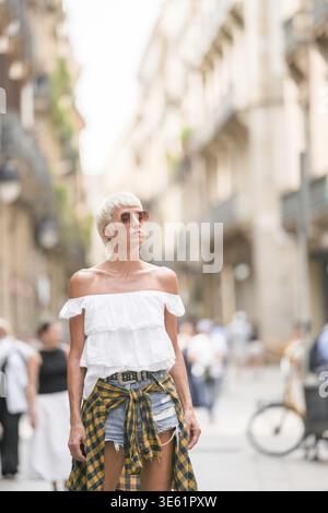 Una donna che indossa una camicia bianca e pantaloncini a quadri cammina lungo una strada. Indossa occhiali da sole e una cintura nera Foto Stock