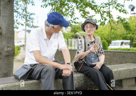 Un uomo e una donna più anziani sono seduti su una panchina in un parco. L'uomo indossa un cappello blu e la donna indossa un cappello grigio. Stanno parlando con ognuno di loro Foto Stock
