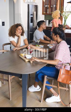 Amici diversi che si riuniscono al tavolo della cucina di casa, donna in maglietta a righe che cerca i cracker Foto Stock