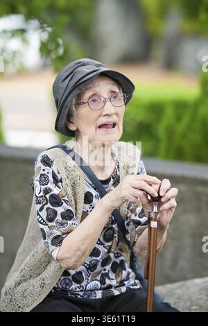 Una donna anziana che indossa un cappello e degli occhiali siede su una panchina con un bastone. Lei è di umore contemplativo Foto Stock