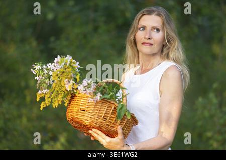 Una donna si erge su un vivace sfondo verde, tenendo in mano un cesto intrecciato pieno di fiori colorati. Sunlight mette in risalto le sue caratteristiche mentre le piace Foto Stock