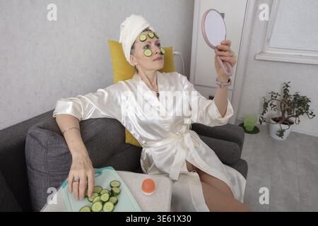 Un'anziana vestita con abiti bianchi, si siede comodamente su un divano grigio, applicando fette di cetriolo sul viso quando guarda in un piccolo specchio. Un tr Foto Stock