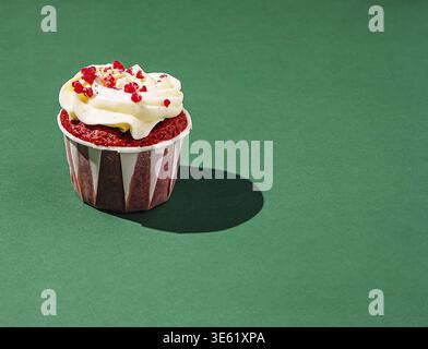 Il cibo, Un cupcake di velluto rosso è collocato su uno sfondo verde vivace. Ricoperto da una glassa di formaggio cremoso liscio e da spruzzi colorati, crea un visu Foto Stock
