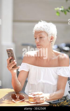 Una donna è seduta a un tavolo con un bicchiere di birra e un cellulare. Sta guardando il telefono mentre tiene il vetro Foto Stock