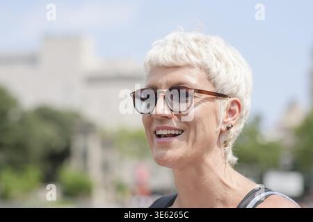 Una donna con capelli biondi corti e occhiali sorride. Indossa una camicia nera e una borsa nera Foto Stock