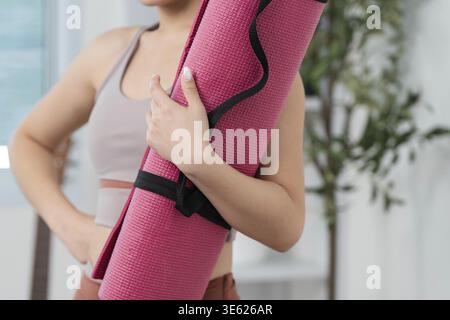 Una giovane donna sicura di sé tiene il suo tappetino da yoga rosa, preparandosi per l'allenamento in un ambiente domestico sereno Foto Stock