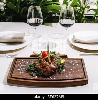 Cibo, Un piatto di agnello splendidamente placcato guarnito con erbe è presentato su un tavolo di legno. Due eleganti bicchieri da vino rosso accompagnano il Foto Stock