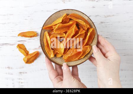 Una donna tiene un piatto con fette di cachi essiccati maturi in un piatto sul tavolo. Uno spuntino sano. Un regalo. Messa a fuoco selettiva, vista dall'alto Foto Stock