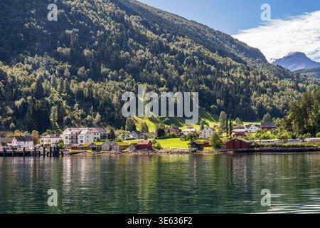 Vista pittoresca di Eidsdal, Norvegia. Case colorate e capannoni con tetti in erba fiancheggiano le calme acque del Norddalsfjord. Una lussureggiante torre di montagna boscosa sopra di te Foto Stock