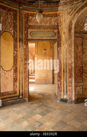 AGLIÈ, ITALIA? OTTOBRE 2025: Vista interna del castello agli è con pareti dipinte ornamentali, marmi invecchiati e porte che conducono ad un corridoio illuminato dal sole, simboleggiando il patrimonio e l'architettura reali italiani. Foto Stock