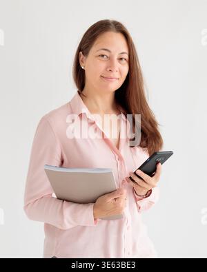 Una donna con lunghi capelli marroni e una camicia rosa chiaro con colletto si staglia su un semplice sfondo bianco, tenendo uno smartphone nero nella mano destra e. Foto Stock
