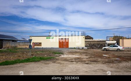 Piccolo edificio beige con porta rossa sotto il cielo blu. Terreno asciutto circonda la struttura con erba sparsa. Auto parcheggiata vicino all'edificio. La scena sembra silenziosa e a Foto Stock