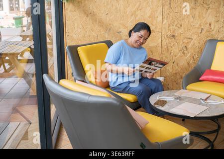 Una donna è seduta in un bar guardando il menu. Si siede su una comoda sedia e si diverte nel caffè con un'atmosfera rilassata. Foto Stock