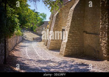 Una strada acciottolata illuminata dal sole si snoda attraverso una storica cittadina europea, fiancheggiata da antiche mura di pietra e contrafforti architettonici sulla destra e lussureggiante Foto Stock