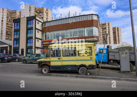Vecchio furgone giallo parcheggiato accanto al camion blu vicino a edifici moderni. Il veicolo in condizioni atmosferiche mostra segni di usura. Ambientazione urbana con auto parcheggiate e apa multipiano Foto Stock