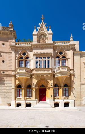 Vista soleggiata di Casa Gourgion a Mdina, Malta, con facciata in pietra neogotica, finestre ad arco a punta, balcone e porta d'ingresso rossa su una superficie pavimentata Foto Stock