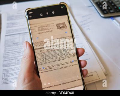 Kirov, Russia - 18 marzo 2026: Una persona scansiona una bolletta con uno smartphone mentre è circondata da documenti cartacei e da una calcolatrice per la pianificazione del budget. Foto Stock