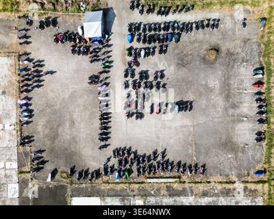 Un grande gruppo di persone si trova in un parcheggio. Le persone sono allineate in file, e ci sono molti ombrelli sparsi per tutta la scena. SCE Foto Stock