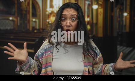 Donna con le mani alzate e le palme aperte, che indossa occhiali e cardigan a motivi geometrici negli interni ornati dell'edificio della chiesa, gesti mentre parla; confusione Foto Stock