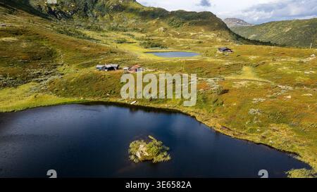 Una splendida vista aerea mostra un lago blu scuro con una piccola isola, circondata dalla tundra norvegese. Cabine tradizionali e un secondo lago sono visibili Foto Stock