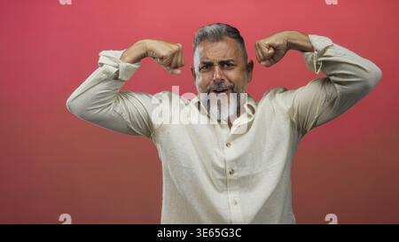 Uomo che si flette bicipite e si afferra mentre solleva le braccia in studio contro il gradiente rosa, la barba grigia e la camicia di lino color crema; sicurezza. Foto Stock